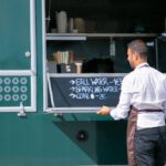 Sales Process - Back view of male seller wearing apron preparing food truck with menu written on board